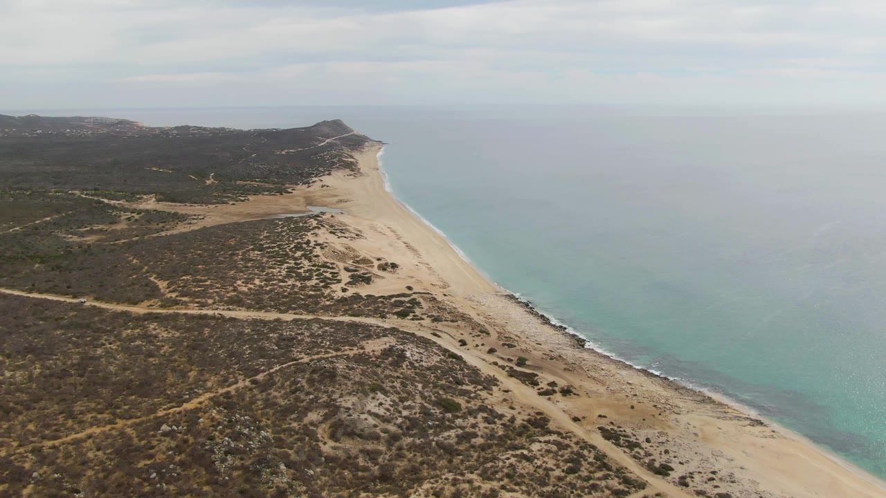 Aerial frame over Punta Gorda Beach, Baja California Sur, with sandy shoreline running diagonally beside turquoise water, scrubland and dirt tracks inland, and no visible people.