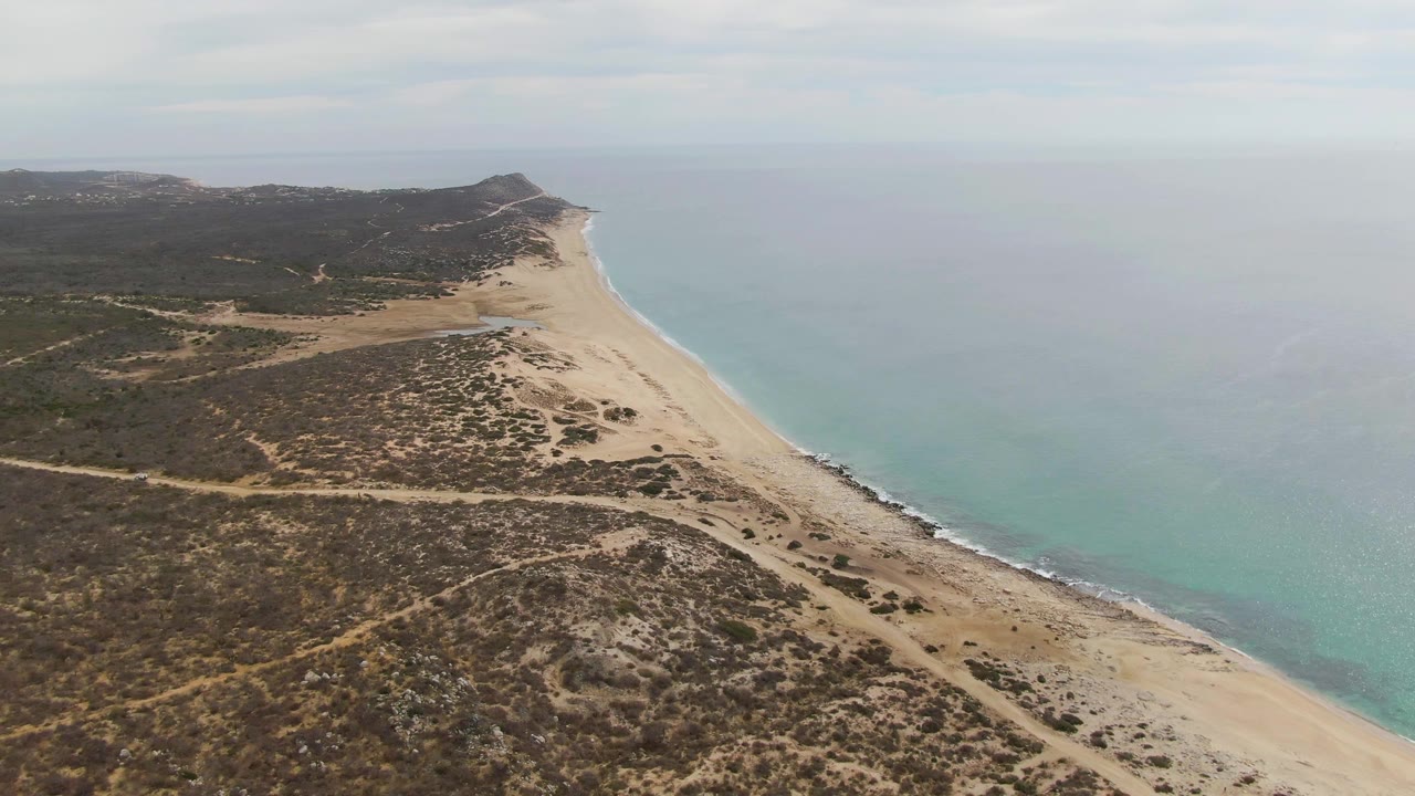 High drone frame over Punta Gorda Beach showing a long diagonal strip of sand, turquoise ocean to the right, scrub-covered dunes to the left, distant hills, and no visible people.