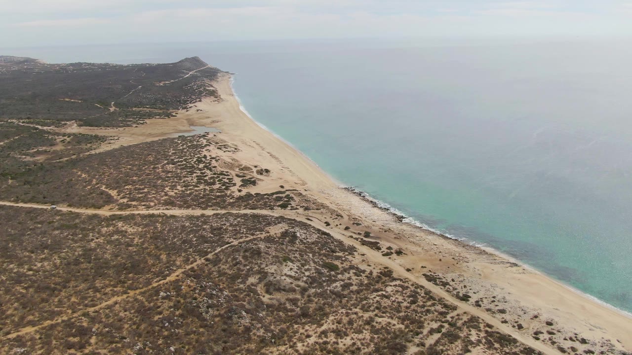 High aerial frame of Punta Gorda Beach, showing scrubby dune terrain and pale tracks beside a long sand strip, with the ocean shifting from turquoise near shore to muted blue offshore and no visible people.