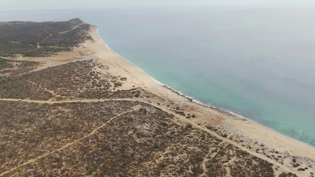 Aerial frame over Punta Gorda Beach BCS showing a sandy shoreline beside turquoise water, dry scrubland and dirt tracks in the foreground, and no visible people.