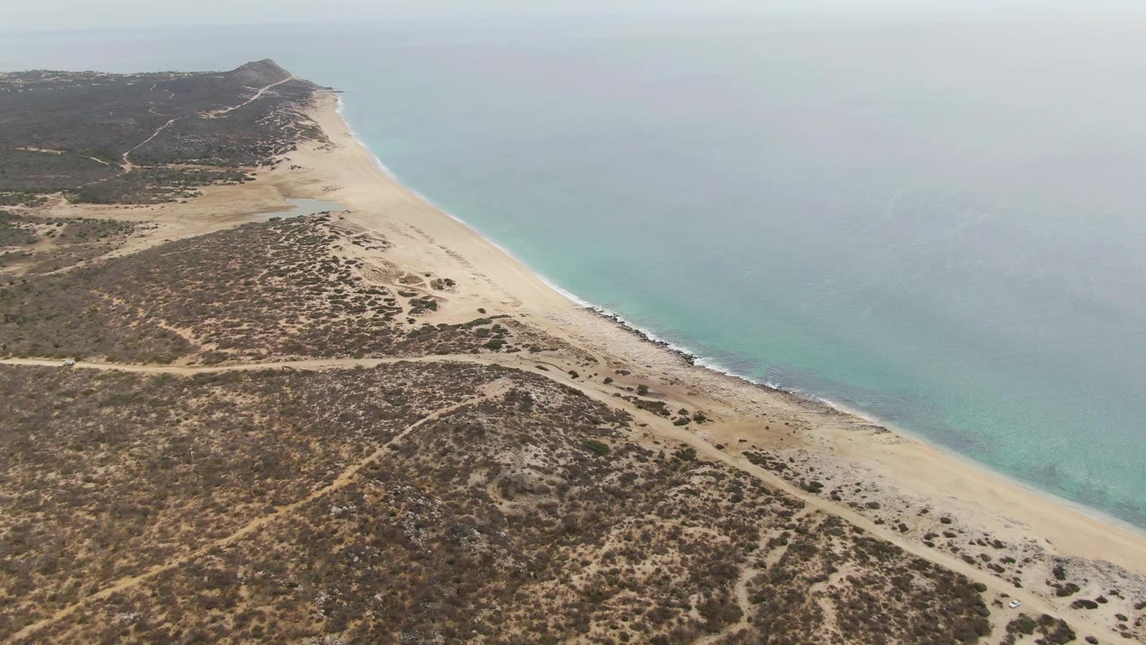 Aerial view of Punta Gorda Beach BCS with the beach curving toward a low headland, dry brown vegetation crossing the foreground, pale surf at the shore, and no visible people.