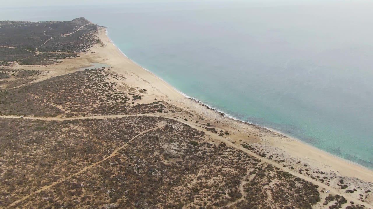 Aerial frame at Punta Gorda Beach BCS showing blue-green ocean on the right, a narrow pale beach, scrub-covered dunes and crossing dirt tracks on the left, with no visible people.