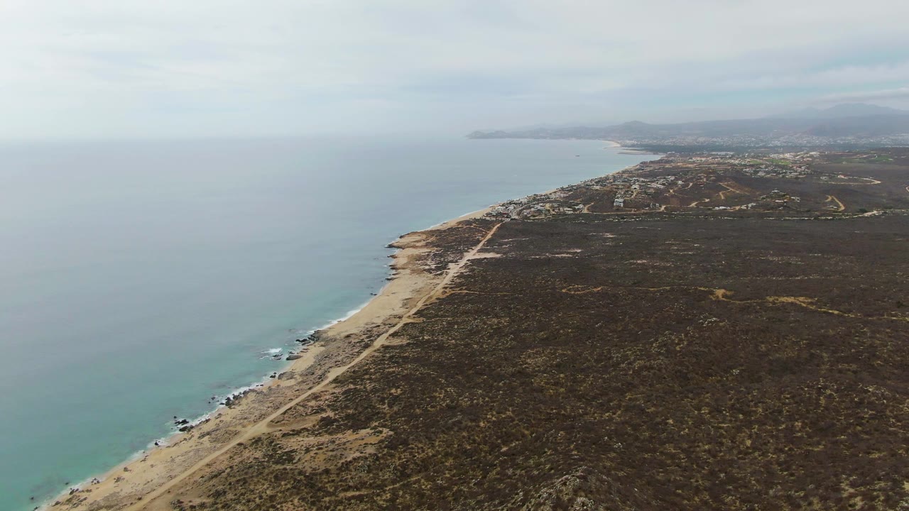Aerial view over Punta Gorda Beach in Baja California Sur, with calm ocean on the left, sandy shore and coastal road below, scrubland to the right, distant hills, and no visible people.
