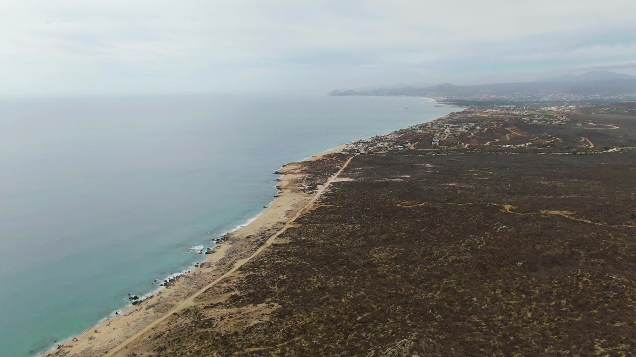 Aerial frame from Punta Gorda Beach with calm water, a pale sandy edge, a road running inland of the beach, broad scrub-covered ground, faint distant development, hills, and no visible people.