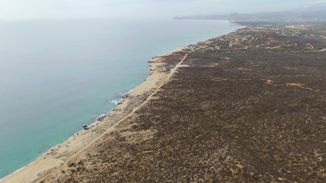 Aerial view at Punta Gorda Beach, Baja California Sur, with turquoise water on the left, a sandy beach and pale road along dry scrubland, and no visible people.