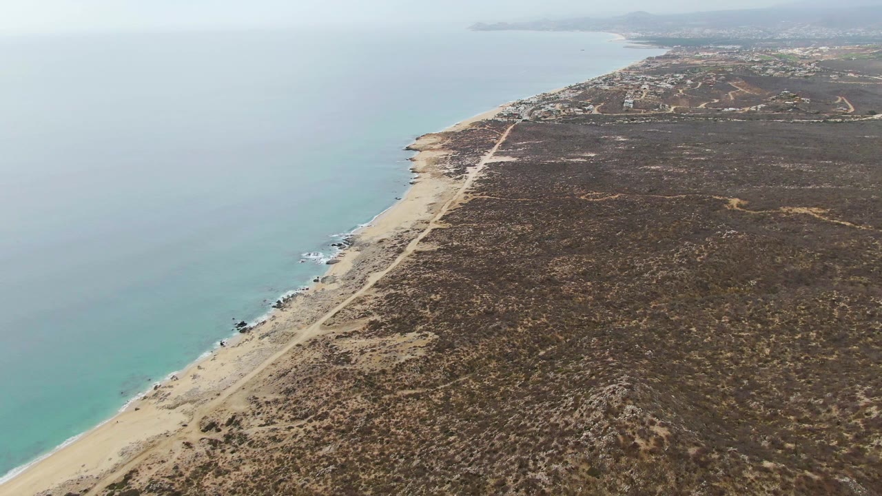 Aerial frame over Punta Gorda Beach showing the beach road closer to center, pale surf along the sand, dry brown vegetation inland, and no visible people.