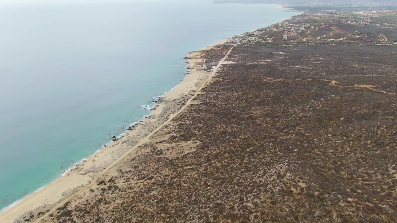 Drone view at Punta Gorda Beach with turquoise ocean and small wave breaks beside the sand, a straight coastal road through dry terrain, distant buildings, and no visible people.