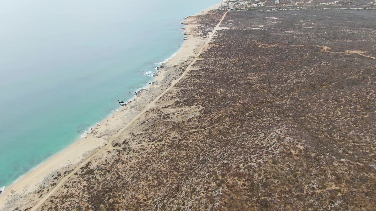 Aerial view at Punta Gorda Beach BCS showing the shoreline running diagonally through frame, turquoise ocean to the left, rough dry terrain to the right, and no visible people.