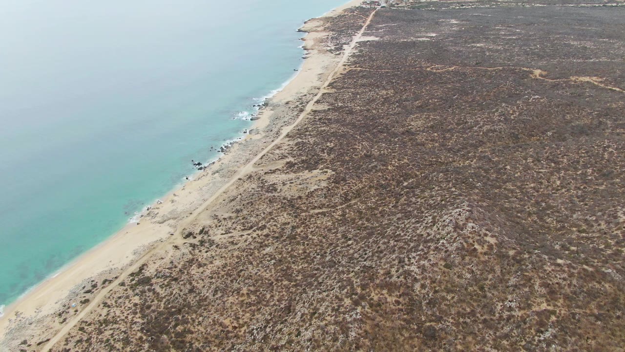 Punta Gorda Beach BCS seen from above with the sandy beach and road parallel to turquoise water, dark scrubland inland, small surf marks near rocks, and no visible people.
