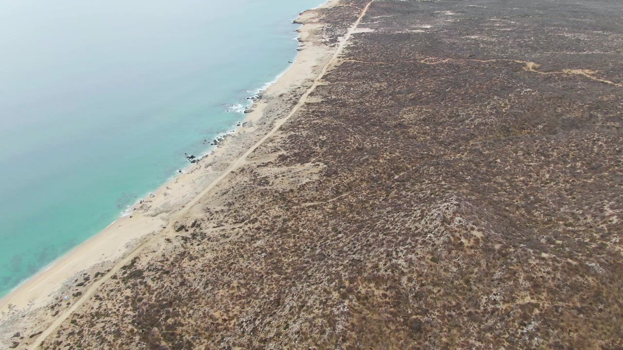 A later aerial frame over Punta Gorda Beach BCS with more open turquoise water on the left edge, the sandy strip curving through the lower frame, dry inland brush, and no visible people.