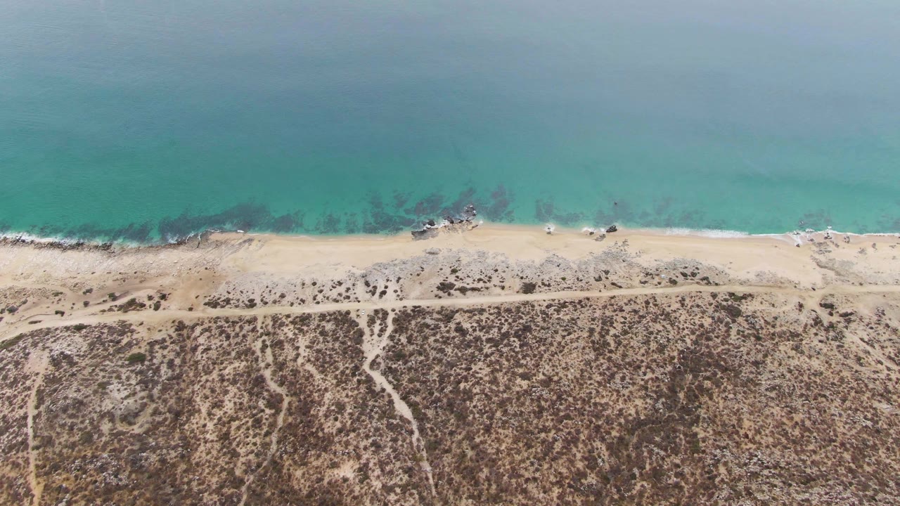 Aerial frame over Punta Gorda Beach showing a thin white surf line, pale sand, a parallel dirt road, and brown arid ground with no visible people.