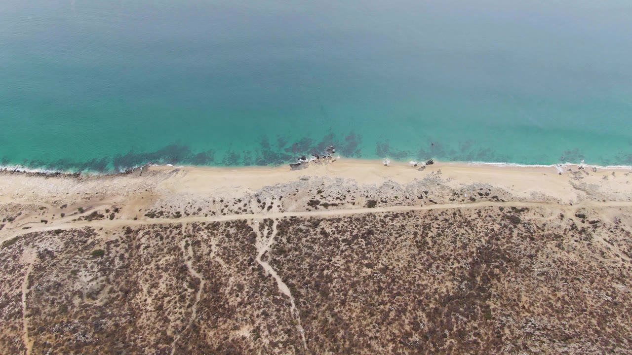 Drone view at Punta Gorda Beach with turquoise ocean across the top, small waves on the sand, and dry Baja California Sur terrain below, with no visible people.