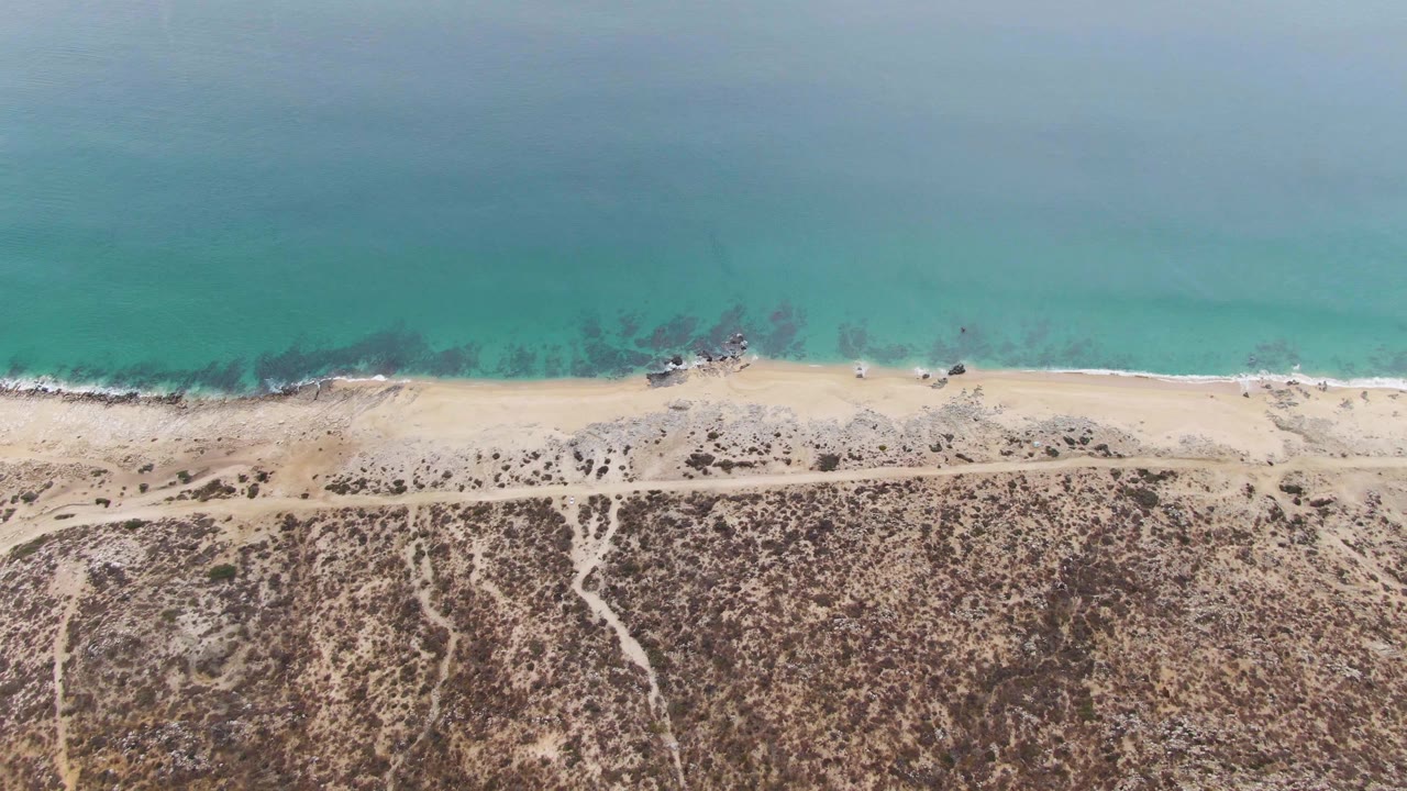 Overhead Punta Gorda Beach frame showing muted turquoise sea, a light sand band, scattered rocks near the waterline, and empty arid inland terrain with no visible people.