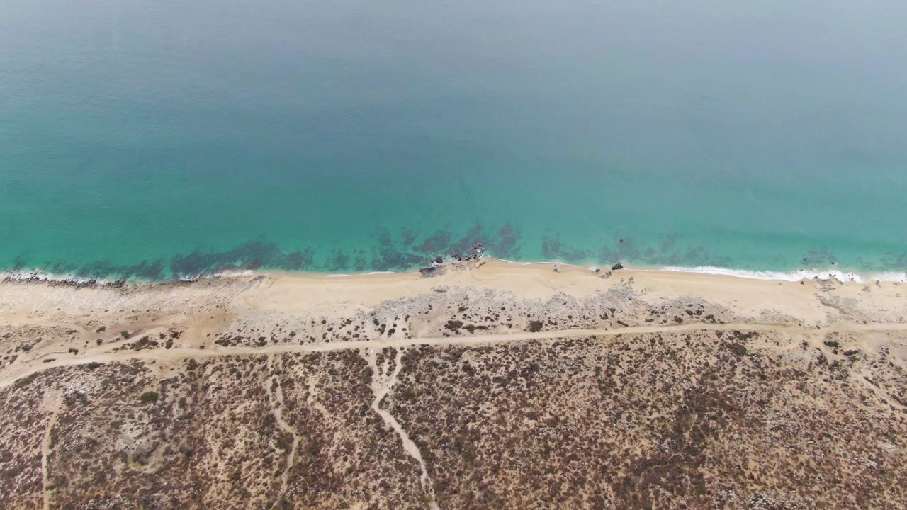 Overhead view of Punta Gorda Beach with turquoise water meeting pale sand, a narrow path behind the shore, dry scrubland below, and no visible people.