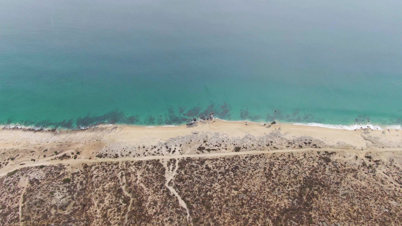 Aerial coastline with small waves, sandy shore, and scrub-covered terrain.