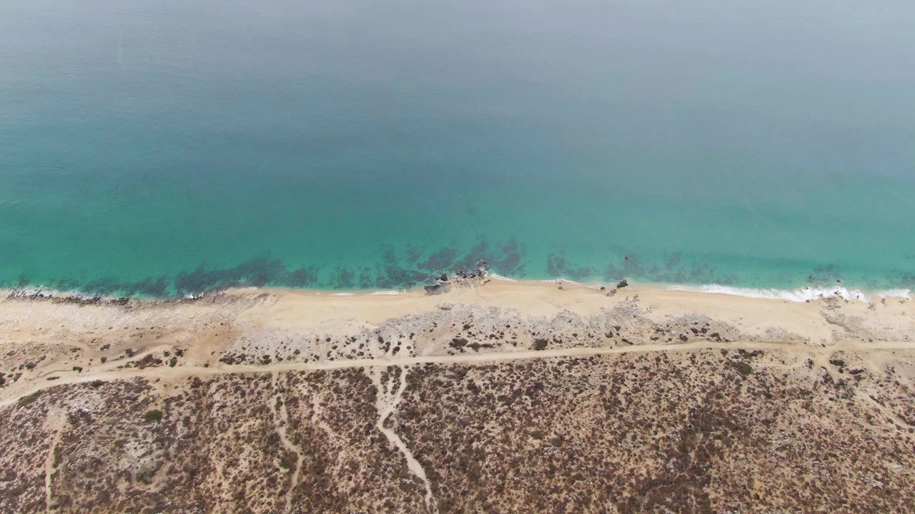 Top-down view of Punta Gorda Beach with a pale sandy strip between turquoise ocean and dry vegetation, plus a thin coastal path and no visible people.