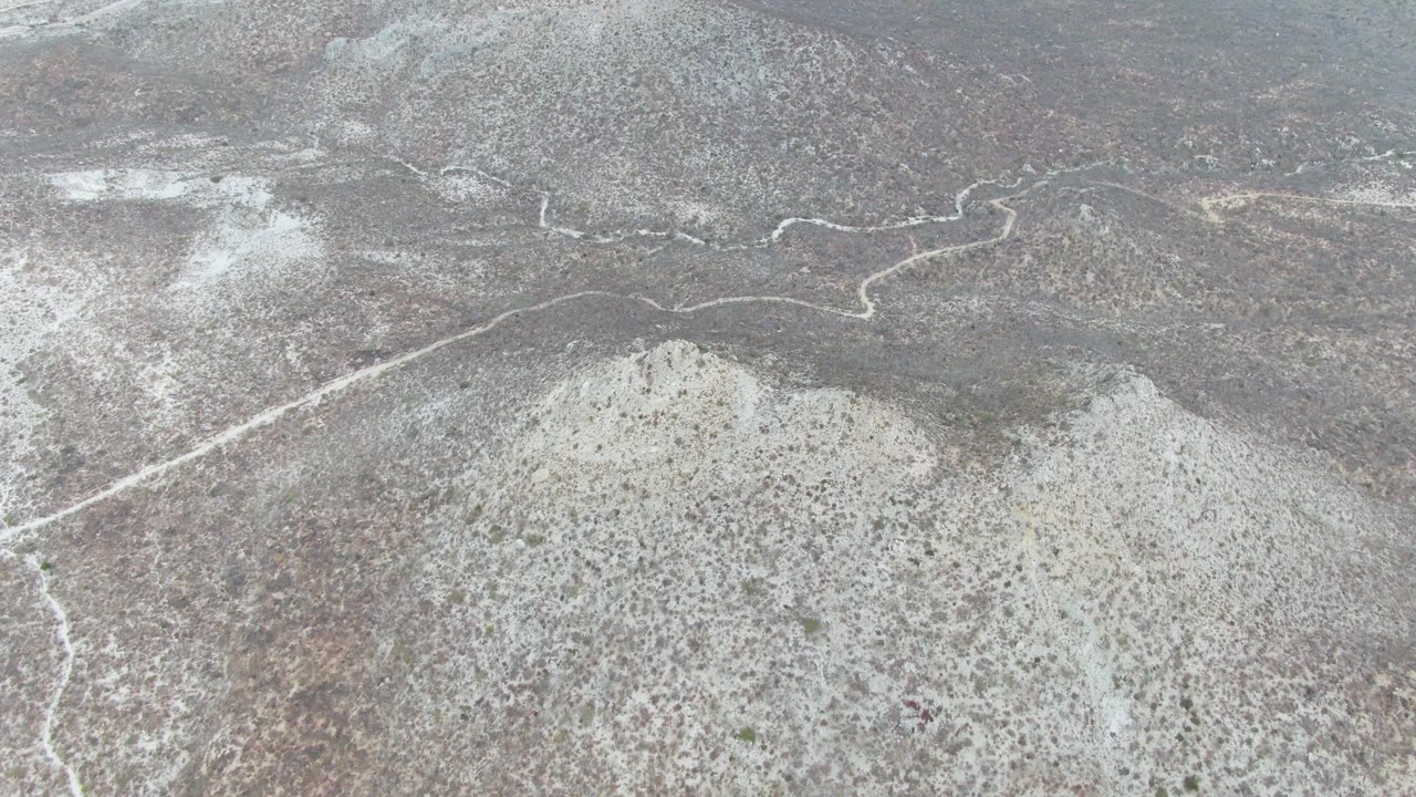 Aerial frame over Punta Gorda Beach, Baja California Sur, showing a pale winding trail crossing gray-brown rocky desert with sparse scrub and no visible people.