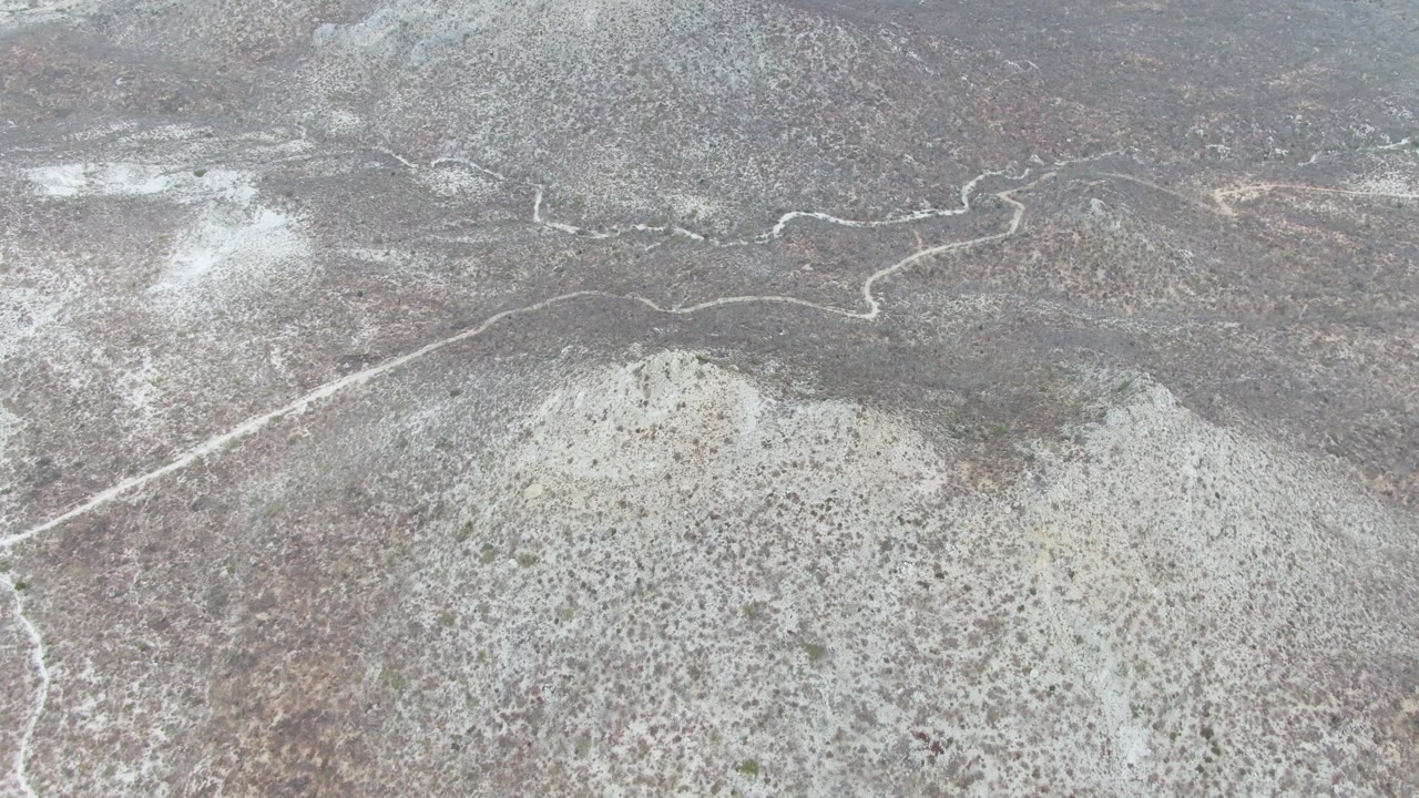 Aerial view of Punta Gorda Beach terrain where a thin dirt trail winds past pale dry washes and scrub-covered gray-brown slopes, with no visible people.