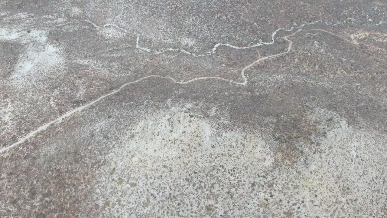 Overhead view of arid Punta Gorda Beach terrain in Baja California Sur, with a pale winding path near the top of frame, mottled brown gray ground, sparse vegetation, and no visible people.