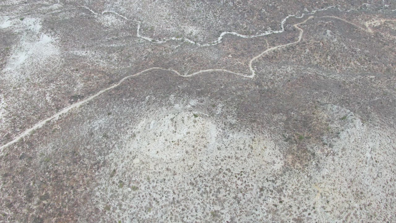 Overhead Punta Gorda Beach frame with dry brown and gray terrain, a pale diagonal track entering from the lower left toward the winding path, patchy scrub, and no visible people.