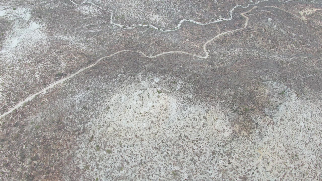 Overhead view of Punta Gorda Beach desert terrain, with the white path curving near the top, mottled brown gray ground filling the frame, pale scrub patches, and no visible people.