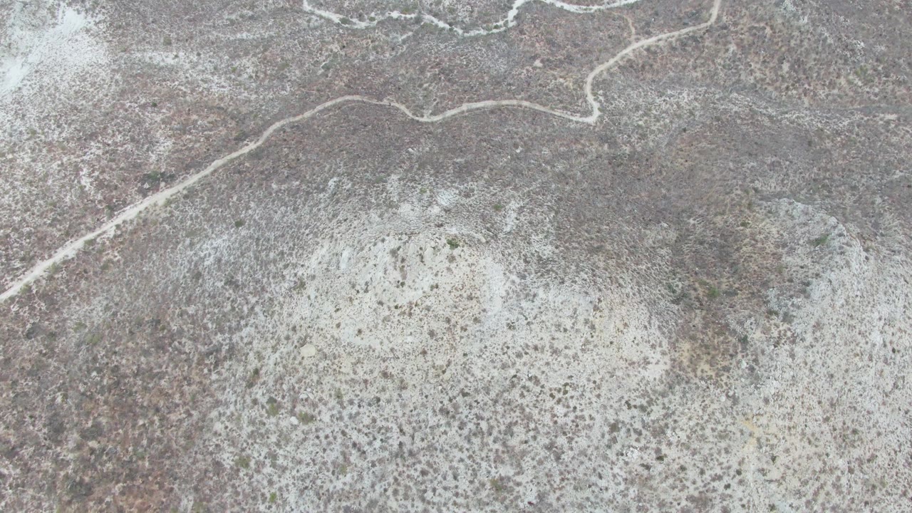 Aerial view of a pale dirt road curving across rocky gray-brown terrain near Punta Gorda Beach, Baja California Sur, with sparse scrub and no visible people.