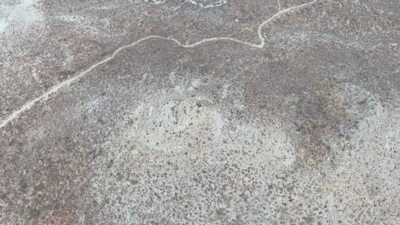 Aerial frame of the dirt road bending through dry gray and brown terrain at Punta Gorda Beach, Baja California Sur, with chalky soil patches and no visible people.