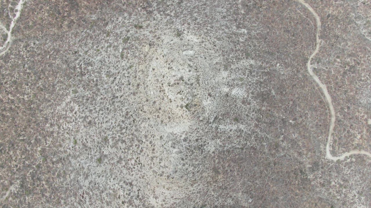Top-down Punta Gorda Beach frame with pale foam patches broken across gray and tan dry terrain, with no visible people or structures.