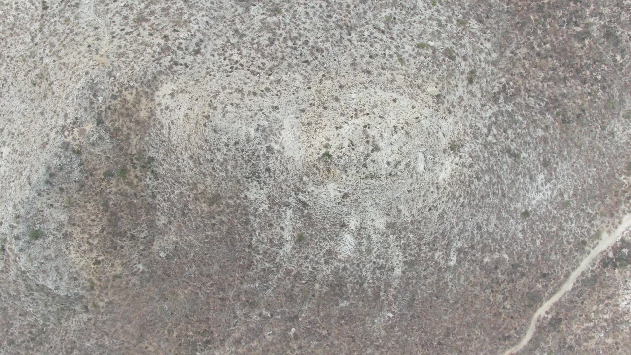 Aerial frame of dry rocky ground at Punta Gorda Beach, Baja California Sur, where a small dark animal shape is visible against sparse scrub, with no visible people.