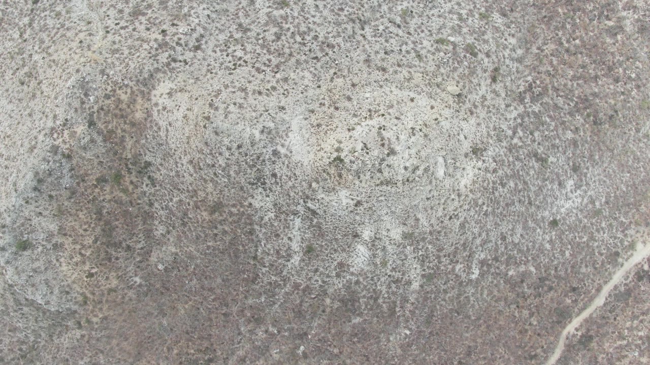 Aerial frame over Punta Gorda Beach BCS arid terrain showing pale rocky patches, sparse scrub, a small light-colored vehicle, and no visible people.
