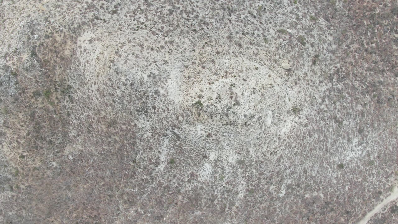 Aerial view over Punta Gorda Beach arid terrain, showing scrub-speckled earth and a faint diagonal dirt track along the lower edge with no visible people.