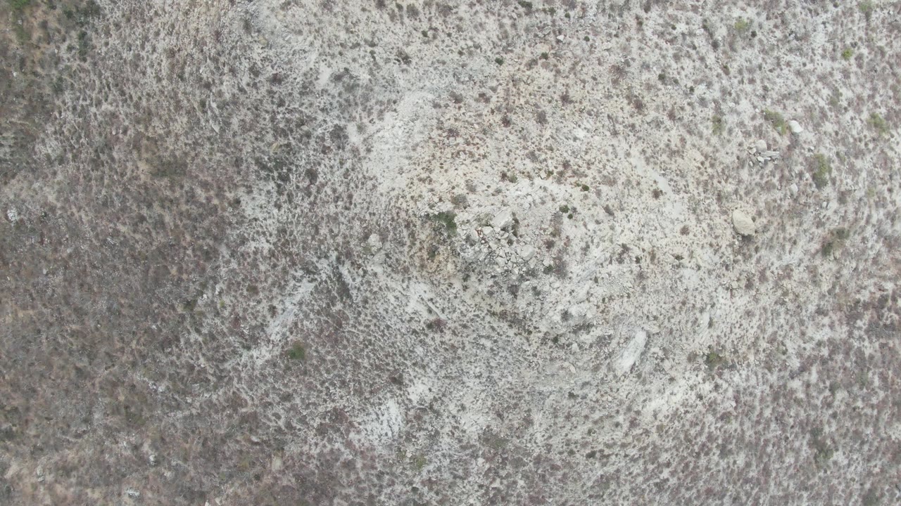 Overhead frame from Punta Gorda Beach, Baja California Sur, showing pale sandy ground, patchy dark scrub, and small rocks with no visible people.