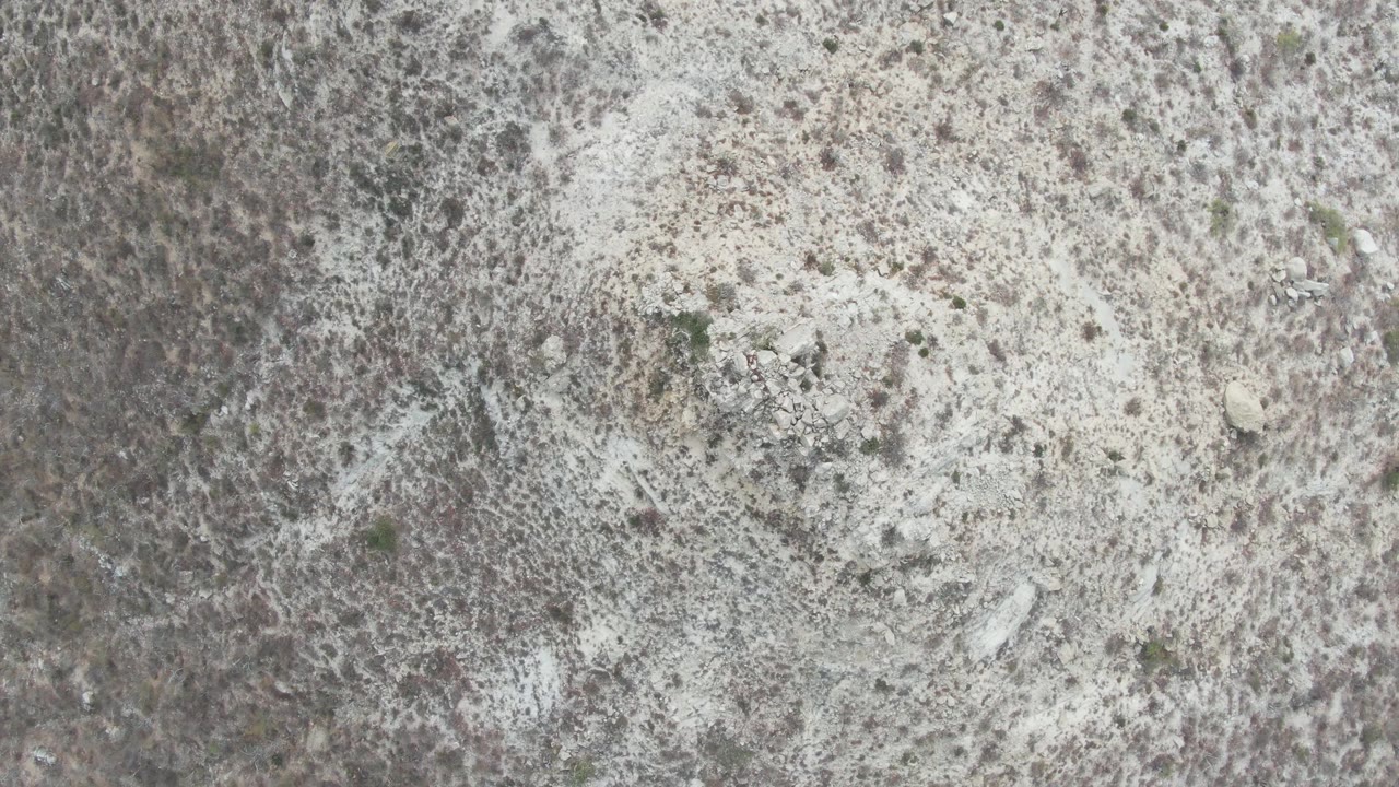 Aerial frame from Punta Gorda Beach showing dry sandy surface, darker brush clusters, and scattered stones, with no visible people.