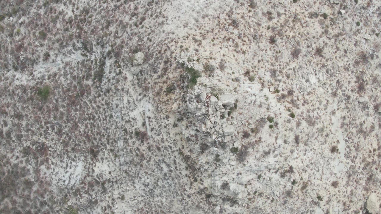 Mid-clip overhead frame from Punta Gorda Beach with pale desert surface, dense mottled scrub areas, small rocks, and no visible people.
