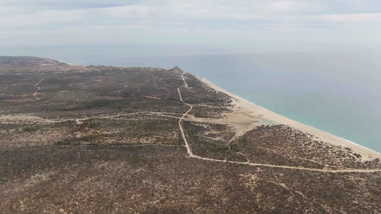 Aerial view at 0:01 over Punta Gorda Beach with branching dirt tracks across brown scrubland, a pale beach strip on the right, calm ocean beyond it, and no visible people.