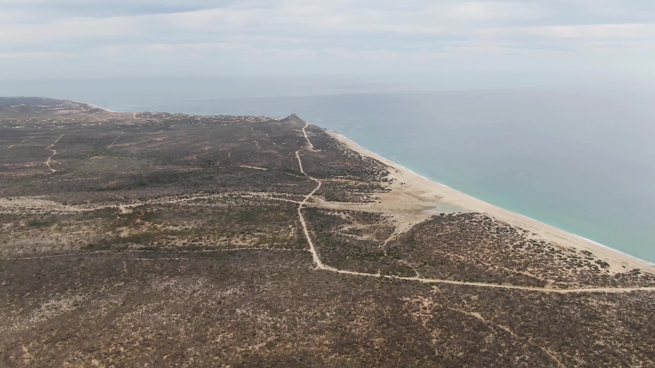 Mid-clip aerial frame from Punta Gorda Beach showing the beach curve, pale blue-green ocean, scrub-covered terrain, crossing paths, distant landform, overcast sky, and no visible people.