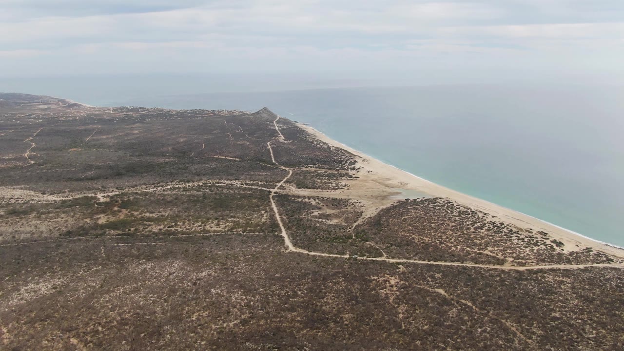 Aerial frame over Punta Gorda Beach, Baja California Sur, with intersecting dirt roads through dry scrubland, a sandy shoreline at right, pale blue ocean beyond, and no visible people.