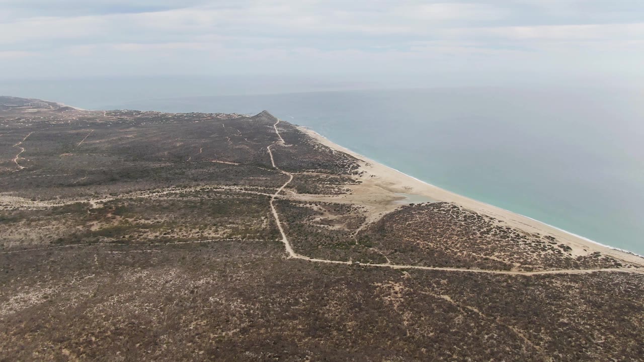 A high drone-like view shows Punta Gorda Beach with pale dirt tracks running toward the coast, dry brown vegetation in the foreground, a sand strip at right, ocean water, and no visible people.