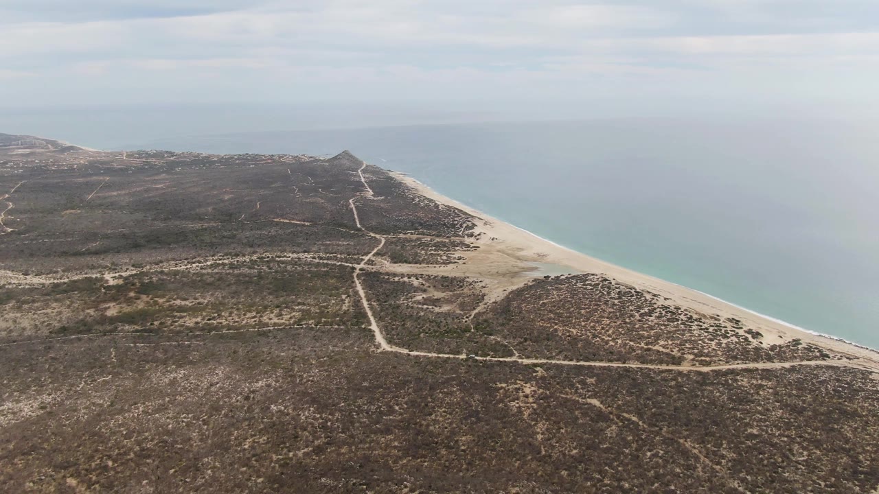 The aerial composition at Punta Gorda Beach places branching dirt roads across dry coastal terrain, the sandy beach along the right edge, calm blue ocean in the distance, and no visible people.