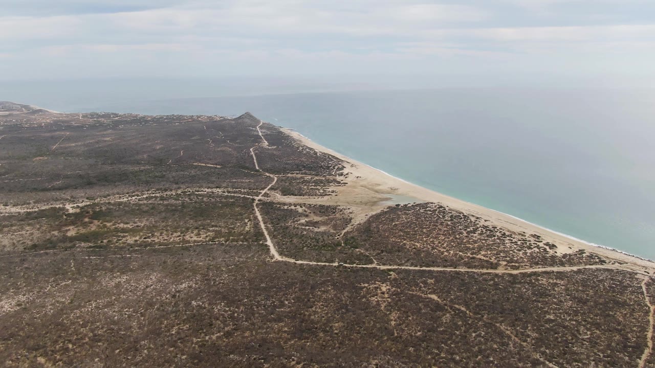 Wide aerial view of coastal scrubland roads beside a long beach and pale ocean.