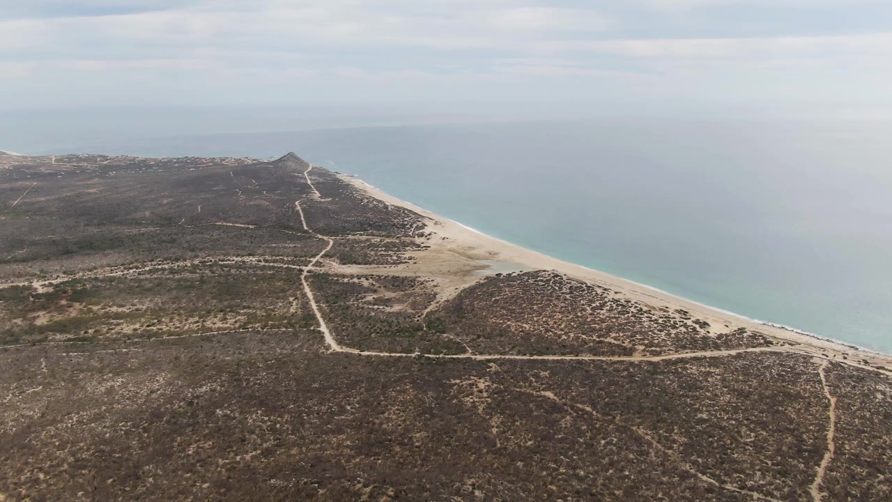 Opening aerial frame over Punta Gorda Beach BCS, Mexico, with a sandy beach curving beside calm turquoise water, dry scrubland, pale paths, and no visible people.