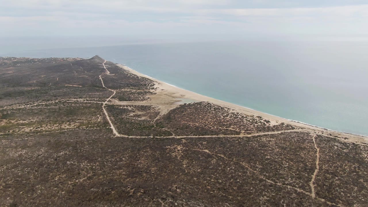 Wide aerial of scrub-covered coastal land, beach, and smooth turquoise water.