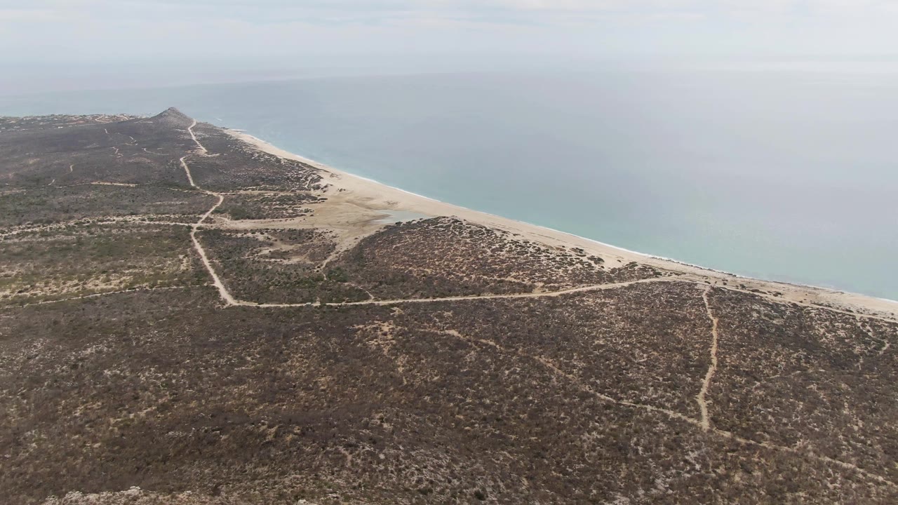 Aerial frame at Punta Gorda Beach, Baja California Sur, showing scrub-covered dry terrain crossed by dirt paths, a pale sandy beach, calm blue ocean, and no visible people.