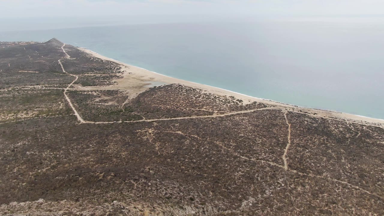 Drone-height frame of Punta Gorda Beach showing a dry headland with pale tracks, a narrow sand strip, calm ocean water, and no visible people.