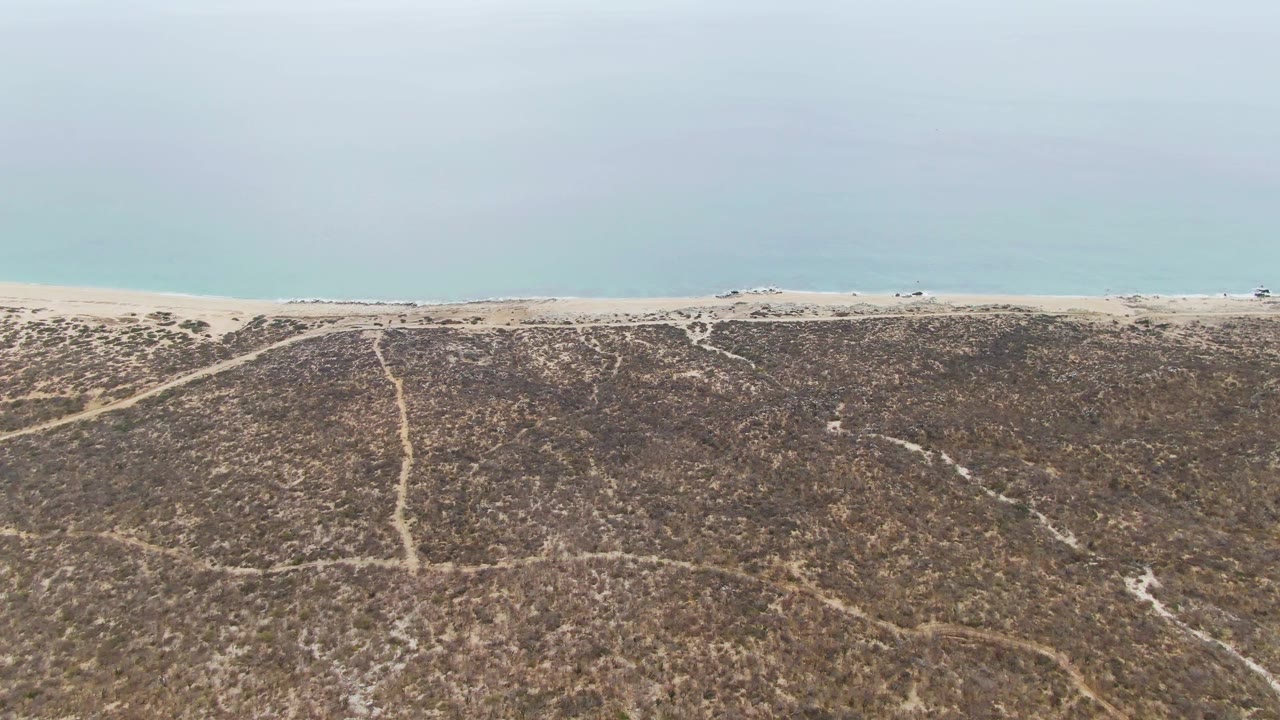 Punta Gorda Beach BCS appears from above with dry brown-green dunes, a central path running toward the sand, a flat beach band, and calm pale water behind it.