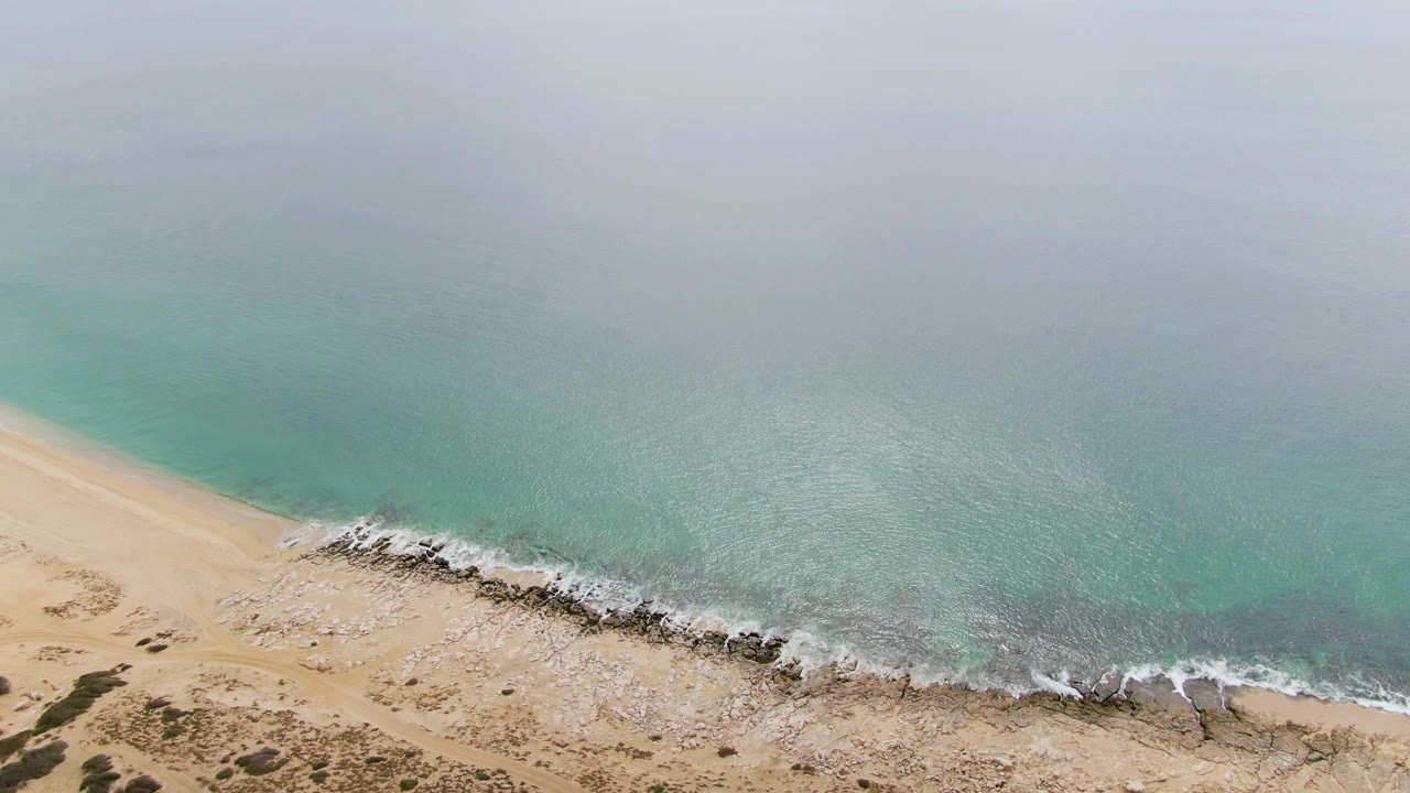 Aerial frame at Punta Gorda Beach showing a diagonal sandy shoreline, scattered rocks in the surf, soft gray light, and no visible people.