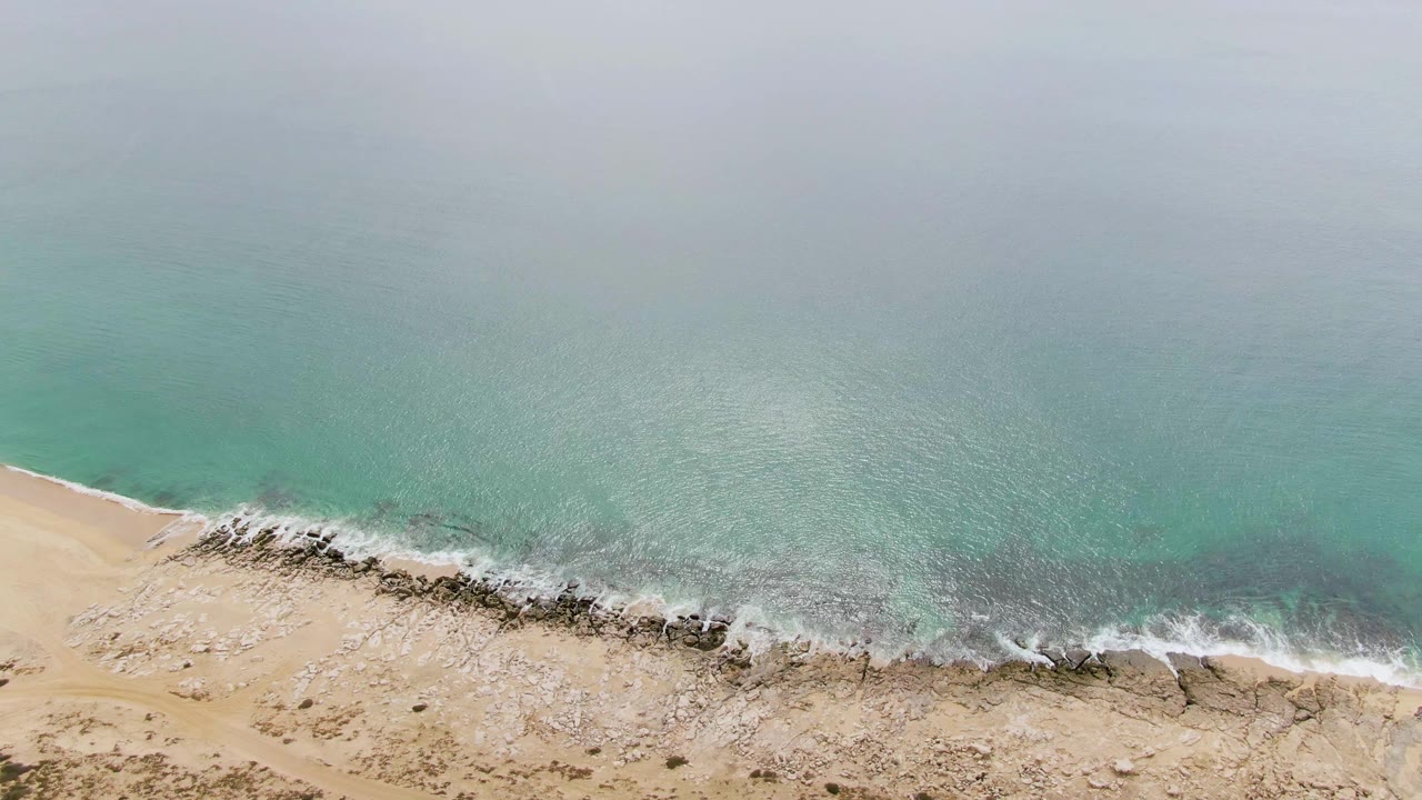 A later aerial frame at Punta Gorda Beach shows calm turquoise water, a narrow band of foam along rocks, pale sand, and no visible people.