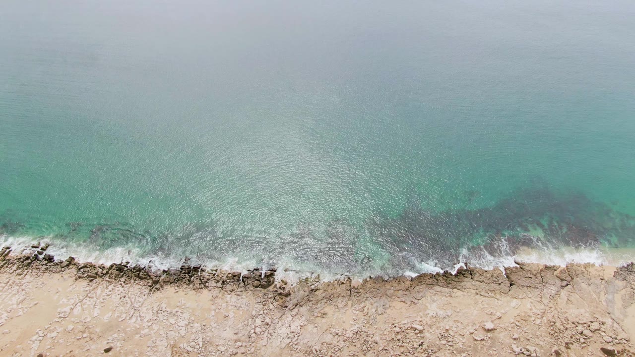 Overhead view of turquoise water breaking into white foam against the rocky sandy shoreline at Punta Gorda Beach, Baja California Sur, with no visible people.