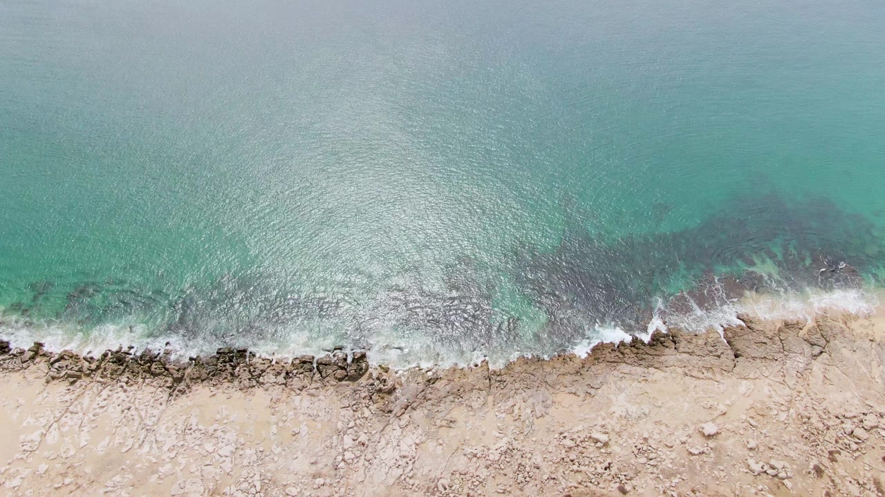 Overhead Punta Gorda Beach view with a wider patch of white surf foam spreading over dark rocks beside pale sand, with no visible people.