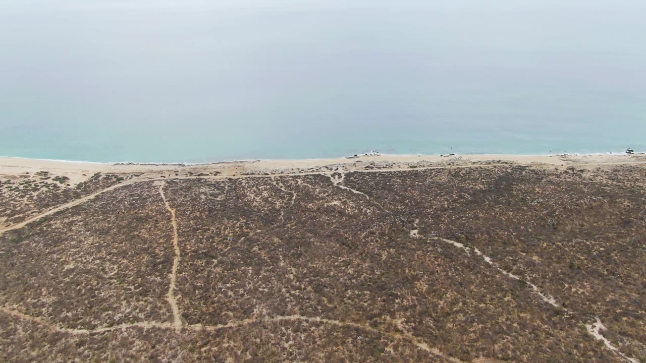 Aerial view of scrub dunes, sandy shoreline, small vehicles, and calm ocean.